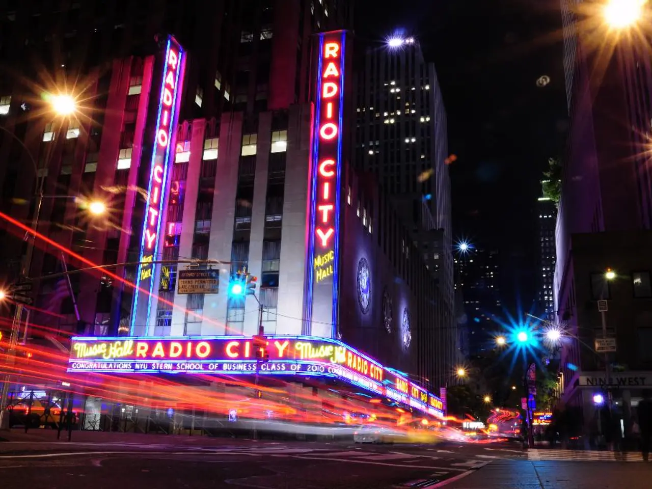 In this image there are buildings and street lights and a few people walking on the road.