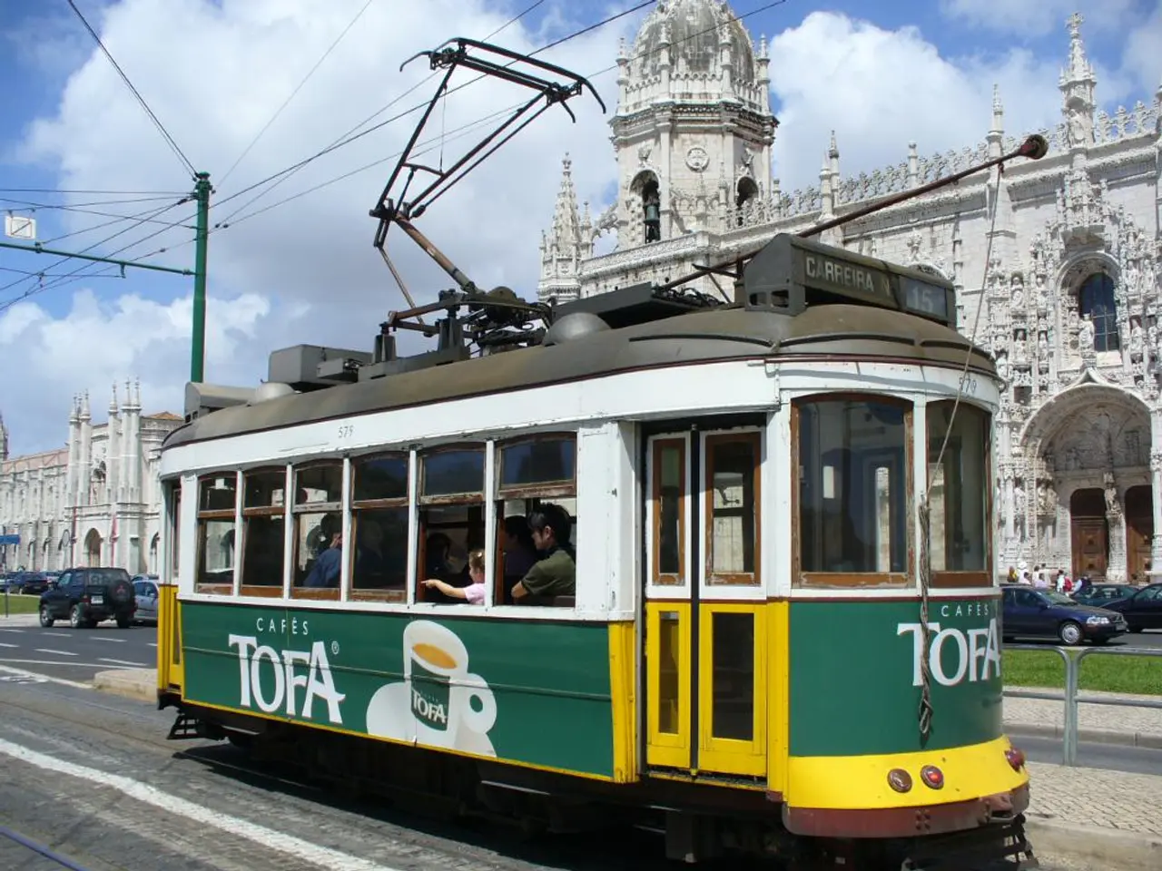 In this image we can see a tram. Inside the tram there are people. In the back we can see vehicles...