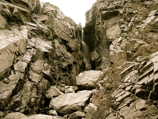 In the image there is a waterfall in the middle of two mountains and above its sky.