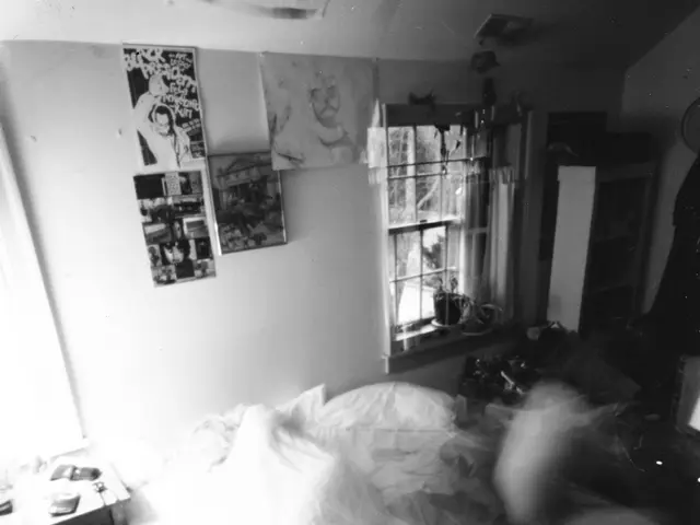 This image consists of a room which contain a bed, a table to the left, a few posts on the wall and...