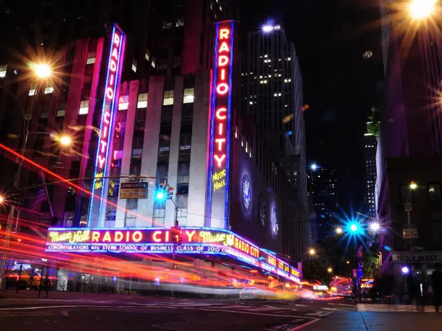 In this image there are buildings and street lights and a few people walking on the road.