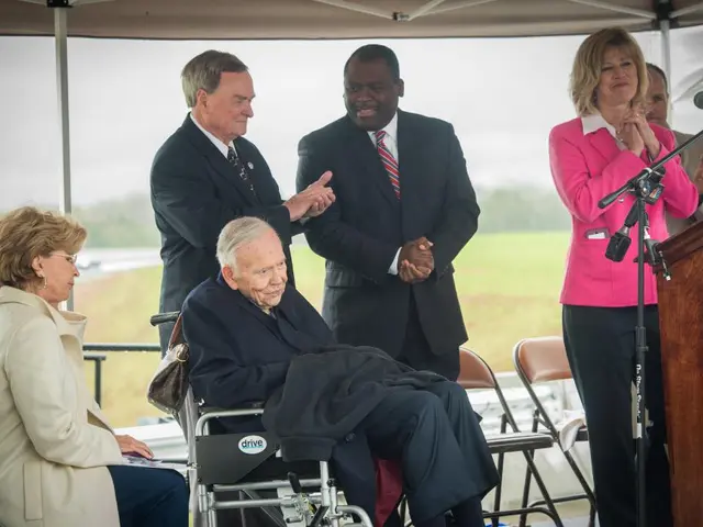 At the bottom of the image two persons are sitting. Behind them few people are standing. Behind...